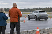 Synthetic editorial illustration. Two engineers viewed from behind hold blank clipboards beside a wet test track. A plain grey car drives among orange cones under an overcast sky with a low treeline beyond.