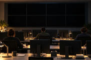 Synthetic editorial illustration. Three operators viewed from behind sit in a dim control room lit by warm desk lamps. Plain monitor backs and fully dark wall screens rise above desks with mugs, plants and blank notebooks.