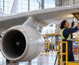 Synthetic editorial illustration. A technician on a platform inspects an outboard wing hatch; the fuselage, wing root and engine suspended below the wing by a pylon are visible as separate structures.