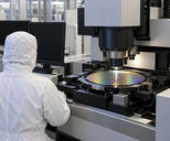 Synthetic editorial illustration. A hooded technician viewed from behind sits beside a patterned wafer under large inspection optics. Two completely dark monitors and plain metal machine housings surround the stage in a bright cleanroom.