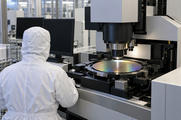 Synthetic editorial illustration. A hooded technician viewed from behind sits beside a patterned wafer under large inspection optics. Two completely dark monitors and plain metal machine housings surround the stage in a bright cleanroom.