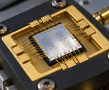 Synthetic editorial illustration. A small silver chip with winding circuit traces sits in a gold-and-black sample holder. Fine gold wire bonds arc between its edges and surrounding contact pads, with worn mounting screws nearby.