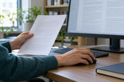 Synthetic editorial illustration. A cropped adult reviews a populated note while using a mouse beside a powered document monitor.