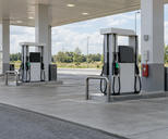 Synthetic editorial illustration. Generic unbranded motor-fuel dispensers and forecourt under a roadside filling-station canopy, with blank/off displays; fuel retail, not natural-gas processing or EV charging.