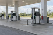 Synthetic editorial illustration. Generic unbranded motor-fuel dispensers and forecourt under a roadside filling-station canopy, with blank/off displays; fuel retail, not natural-gas processing or EV charging.