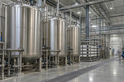 Synthetic editorial illustration. An industrial water-treatment hall. Tall stainless tanks, filtration housings and dense polished pipework line a clean utility space with floor drainage grilles and overhead lighting.
