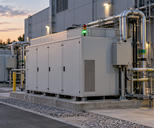 Synthetic editorial illustration. Connected modular utility enclosures with illuminated status lamps stand beside a facility wall at dusk.