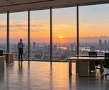 Synthetic editorial illustration. A grey-haired rear-facing person holds a mug beside floor-to-ceiling office windows. Empty desks and chairs frame a broad city view under orange sunrise light.