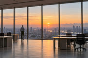 Synthetic editorial illustration. A grey-haired rear-facing person holds a mug beside floor-to-ceiling office windows. Empty desks and chairs frame a broad city view under orange sunrise light.