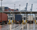 Synthetic editorial illustration. A container-terminal gate. Freight trucks queue beside inspection booths beneath a broad canopy, with stacked containers and cranes behind and red tail lights reflected on wet pavement under an overcast sky.