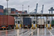 Synthetic editorial illustration. A container-terminal gate. Freight trucks queue beside inspection booths beneath a broad canopy, with stacked containers and cranes behind and red tail lights reflected on wet pavement under an overcast sky.