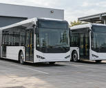 Synthetic editorial illustration. Two generic unbranded electric city buses parked at an anonymous practical depot with charging infrastructure.