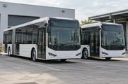 Synthetic editorial illustration. Two generic unbranded electric city buses parked at an anonymous practical depot with charging infrastructure.
