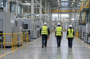 Synthetic editorial illustration. A factory production hall. Three inspectors seen from behind walk along a marked aisle carrying clipboards, wearing bright safety vests and hard hats among machinery and pipework.