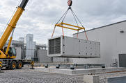 Synthetic editorial illustration. One suspended industrial air-handling module is being positioned beside a service building, without people in the frame.