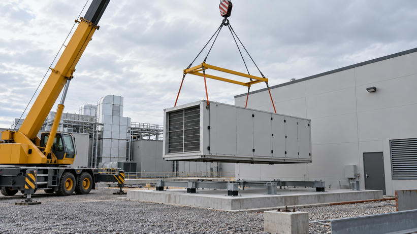 Synthetic editorial illustration. One suspended industrial air-handling module is being positioned beside a service building, without people in the frame.