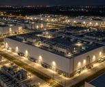 Synthetic editorial illustration. A data-centre campus. An elevated night view shows large windowless buildings, rooftop cooling units, interconnected pipework and brightly lit service roads inside a fenced industrial site.
