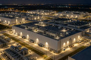 Synthetic editorial illustration. A data-centre campus. An elevated night view shows large windowless buildings, rooftop cooling units, interconnected pipework and brightly lit service roads inside a fenced industrial site.
