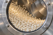 Synthetic editorial illustration. A circular opening in a stainless-steel processing drum frames a bed of small white tablets. The drum wall, a curved internal baffle and the bolted outer rim are visible.