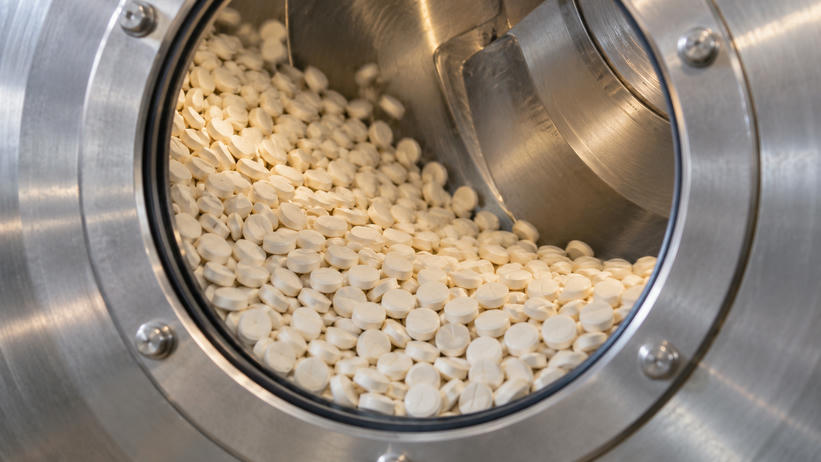 Synthetic editorial illustration. A circular opening in a stainless-steel processing drum frames a bed of small white tablets. The drum wall, a curved internal baffle and the bolted outer rim are visible.