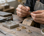 Synthetic editorial illustration. Anonymous jeweller hands working on generic gold-toned rings and a simple chain at a practical craft bench; no recognisable brand design.