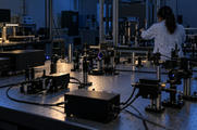 Synthetic editorial illustration. Mounted lenses, mirrors, dark laser housings and cables cover a perforated optical table in subdued blue light. A researcher seen from behind adjusts equipment at the far side.