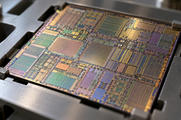 Synthetic editorial illustration. A bare square silicon die rests in a plain metal sample holder. Dense geometric circuitry and rectangular regions reflect muted gold, green and violet light in a shallow-focus macro view.