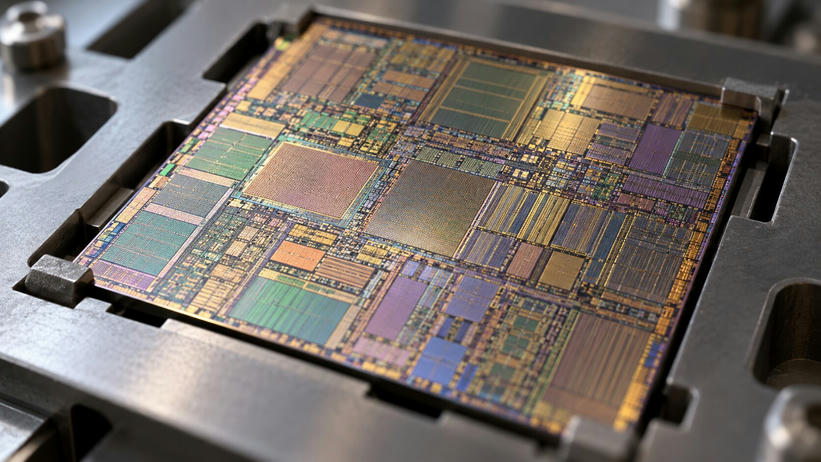 Synthetic editorial illustration. A bare square silicon die rests in a plain metal sample holder. Dense geometric circuitry and rectangular regions reflect muted gold, green and violet light in a shallow-focus macro view.