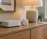 Synthetic editorial illustration. Generic unbranded smart-home gateway and wireless environmental sensor on a domestic sideboard; no real named product.