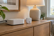 Synthetic editorial illustration. Generic unbranded smart-home gateway and wireless environmental sensor on a domestic sideboard; no real named product.