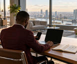 Synthetic editorial illustration. A modern office. A rear-facing person in a burgundy jacket uses a dark laptop and phone at a wooden desk, beside an open notebook with arrows, a mug and a plant; tall windows overlook the city.