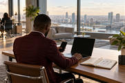 Synthetic editorial illustration. A modern office. A rear-facing person in a burgundy jacket uses a dark laptop and phone at a wooden desk, beside an open notebook with arrows, a mug and a plant; tall windows overlook the city.