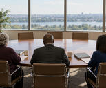 Synthetic editorial illustration. A conference room. Three committee members seen from behind sit along an oval table with plain binders, papers and coffee cups, facing empty chairs and a daylight window view.