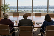 Synthetic editorial illustration. A conference room. Three committee members seen from behind sit along an oval table with plain binders, papers and coffee cups, facing empty chairs and a daylight window view.