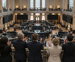 Synthetic editorial illustration. An exchange-hall balcony. A rear-facing group applauds beside a small brass bell above a trading floor of curved desks and dark display panels, framed by tall columns and windows.