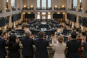 Synthetic editorial illustration. An exchange-hall balcony. A rear-facing group applauds beside a small brass bell above a trading floor of curved desks and dark display panels, framed by tall columns and windows.
