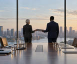 Synthetic editorial illustration. A boardroom overlooks a city through floor-to-ceiling windows. Two rear-facing suited people pass a closed folder between them beside a long table set with plain folders and water glasses.