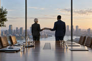 Synthetic editorial illustration. A boardroom overlooks a city through floor-to-ceiling windows. Two rear-facing suited people pass a closed folder between them beside a long table set with plain folders and water glasses.