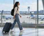 Synthetic editorial illustration. A traveller wearing a backpack pulls one wheeled carry-on through a glass-sided airport corridor.