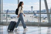 Synthetic editorial illustration. A traveller wearing a backpack pulls one wheeled carry-on through a glass-sided airport corridor.