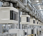 Synthetic editorial illustration. Sealed pale carrier pods with transparent side windows hang in a receding line beneath metal transport rails. Stacked wafer edges are visible inside, above a bright factory with dark equipment displays.