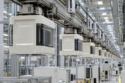 Synthetic editorial illustration. Sealed pale carrier pods with transparent side windows hang in a receding line beneath metal transport rails. Stacked wafer edges are visible inside, above a bright factory with dark equipment displays.