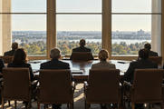 Synthetic editorial illustration. A group of formally dressed people viewed from behind sit around a broad table facing tall bright windows. Plain closed folders rest on the table, with trees and an indistinct city visible outside.