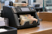 Synthetic editorial illustration. A compact banknote counter feeds an input stack into an output pocket at a currency-service counter.