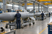 Synthetic editorial illustration. Rows of plain grey finned missile bodies rest on wheeled assembly cradles in a bright industrial hall. Technicians viewed from behind work alongside them beneath yellow overhead cranes.
