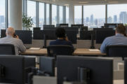 Synthetic editorial illustration. A trading office. Three workers seen from behind hold telephone handsets at a long desk surrounded by rows of monitors facing away, with a soft city view beyond the windows.
