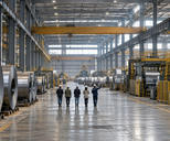 Synthetic editorial illustration. A factory floor contains large metal coils, processing machinery and overhead cranes. Five small rear-facing people in hard hats walk down the broad aisle as one points toward equipment.