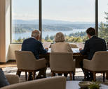Synthetic editorial illustration. An advisory office overlooks water and wooded hills through a large window. A rear-facing older couple and suited adviser sit side by side at a desk holding blank white sheets.