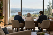 Synthetic editorial illustration. An advisory office overlooks water and wooded hills through a large window. A rear-facing older couple and suited adviser sit side by side at a desk holding blank white sheets.