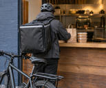 Synthetic editorial illustration. Anonymous bicycle courier seen entirely from behind at a restaurant meal pickup counter, with plain insulated bag and sealed takeaway food containers; no named delivery service.